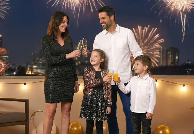 Een gezin viert feest met drankjes op een balkon, terwijl vuurwerk de nachtelijke hemel achter hen verlicht.