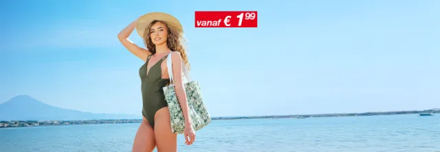 Een vrouw in een groen badpak en een hoed met brede rand staat op het strand en houdt een tas met tropisch design vast tegen een heldere lucht.
