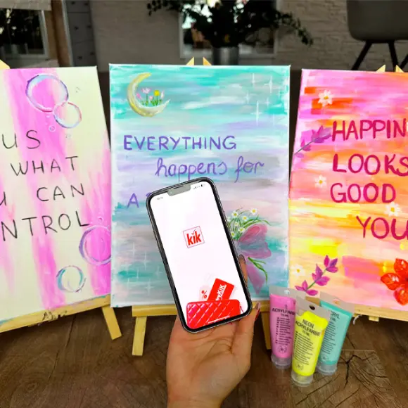 Eén hand houdt een smartphone met de KiK-app vast, daarnaast staan drie kleurrijke schilderijen met motiverende quotes op schildersezels.