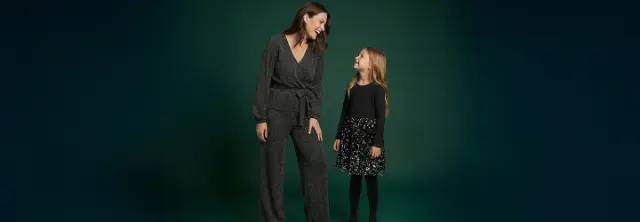 Eine Frau in einem glitzernden schwarzen Overall steht neben einem Mädchen in einem schwarzen Oberteil mit einem Paillettenrock, beide lächeln vor einem dunkelgrünen Hintergrund.