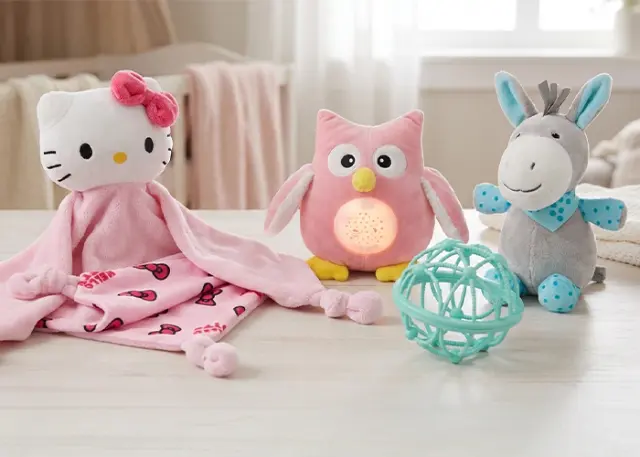 Een knusse opstelling van pluchen speelgoed: een Hello Kitty-dekentje, een roze uil met licht, een grijze ezel en een petrolkleurige bal, allemaal op een houten tafel.