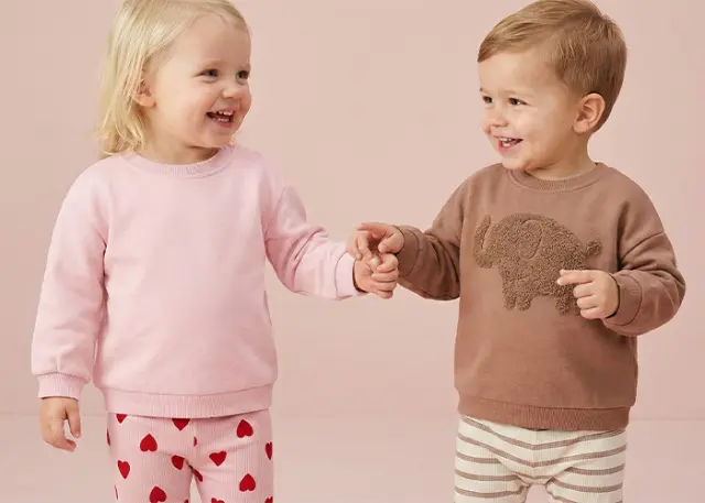 Twee jonge kinderen houden elkaars handen vast, de één draagt een roze trui en een gestreepte broek met hartjesmotief, de ander een bruine trui met een olifantenontwerp.