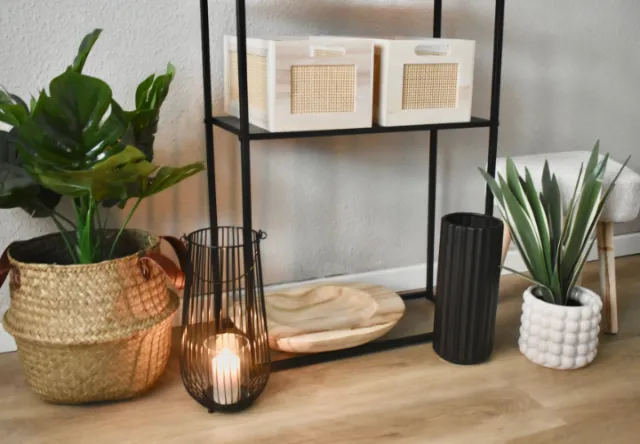 Ein stilvolles Regal präsentiert geflochtene Aufbewahrungsboxen, ein Holztablett, einen Kerzenhalter aus Metall und Topfpflanzen vor einer neutralen Wand.
