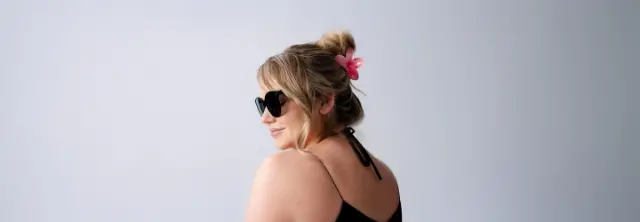 Een vrouw met golvend haar gestyled in een knot versierd met een roze bloem, draagt een zwart topje en poseert van achteren tegen een lichte achtergrond.