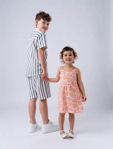 Een jongen in een gestreepte outfit en een meisje in een gebloemde jurk houden elkaars hand vast, beide glimlachend, tegen een effen witte achtergrond.