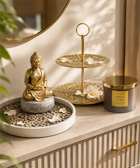 Gouden Boeddhabeeldje op een „Balance”-steen, naast een gouden etagère met bloemblaadjes en een geurkaars „Morning Citrus” op een houten kast.
