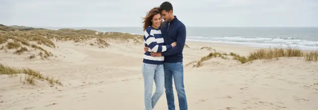 Een stel in casual kleding omarmt elkaar op een zandstrand met duinen en oceaangolven onder een bewolkte lucht.