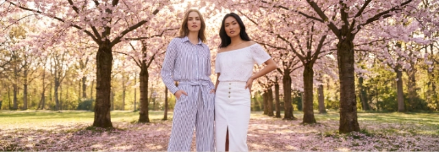 Twee vrouwen presenteren moderne damesmode tussen bloeiende bomen – frisse voorjaarslooks voor stijlvolle outfits