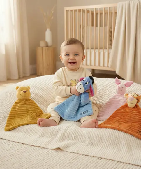 Een baby zit op een zachte deken, omringd door kleurrijke knuffels zoals Iejoor, Knorretje en Teigetje, in een lichte kamer.