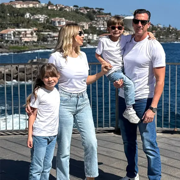 Een gezin van vier poseert samen aan zee, gekleed in bijpassende witte T-shirts en jeans, met een schilderachtig decor van heuvels en water op de achtergrond.