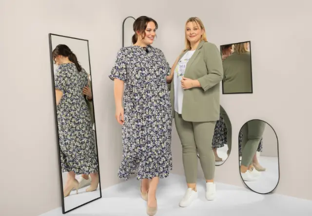Zwei Frauen stehen in einer stilvollen Umgebung mit Spiegeln und präsentieren ein geblümtes Kleid und ein schickes grünes Outfit, das Selbstbewusstsein und Eleganz ausstrahlt.
