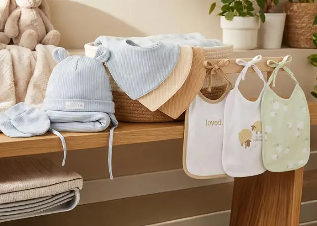 Een knusse presentatie van babybenodigdheden: een blauwe muts, zachte dekens en slabbetjes met schattige designs, gerangschikt op een houten plank.
