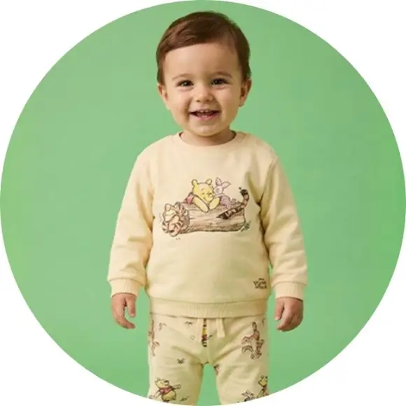 Een peuter staat tegen een groene achtergrond en draagt een gele sweater met een speelse illustratie van cartoonfiguren.