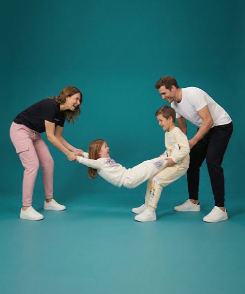 Een gezin speelt speels met elkaar en houdt een kind tussen twee volwassenen; allemaal casual gekleed tegen een turquoise achtergrond.
