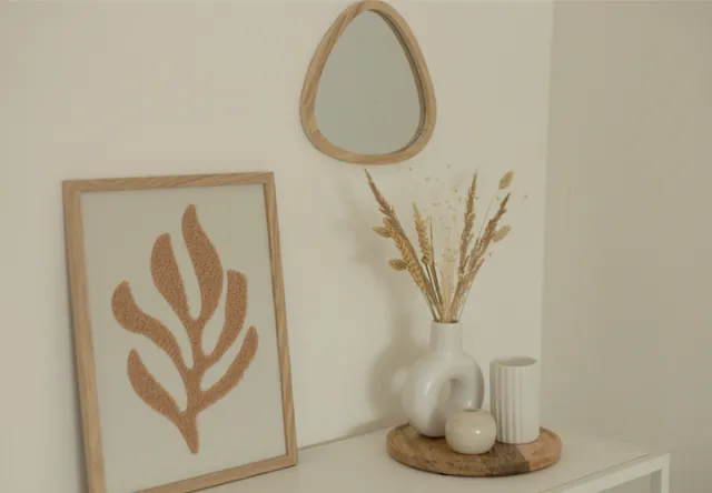 Een minimalistische inrichting met een kleine spiegel, een ingelijste bladkunst en een vaas met gedroogde bloemen op een houten dienblad, tegen een neutrale muur.
