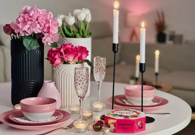 Een prachtig gedekte dinertafel met roze borden, kaarsen, elegant glaswerk en een doos Merci-chocolade, omringd door verse bloemen.