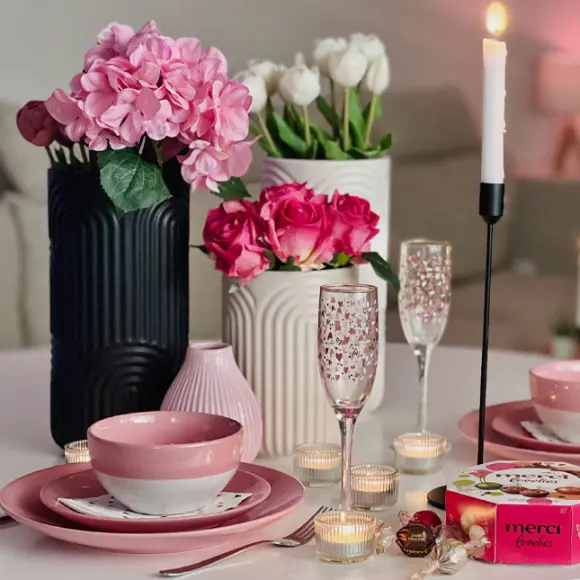 Een prachtig gedekte dinertafel met roze borden, kaarsen, elegant glaswerk en een doos Merci-chocolade, omringd door verse bloemen.