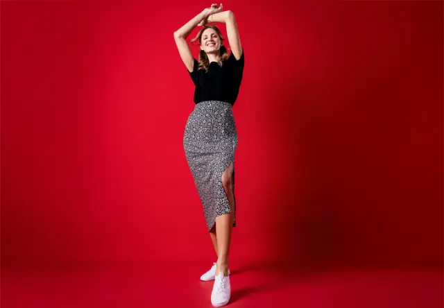 Een vrouw poseert zelfverzekerd voor een felrode achtergrond, gekleed in een zwart topje, een rok met patroon en witte sneakers.
