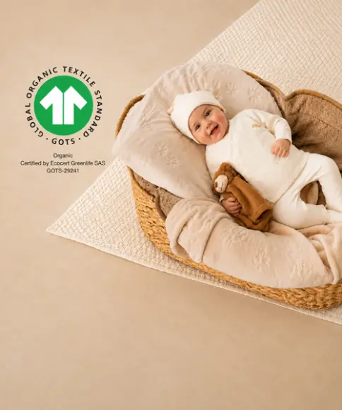 Baby in GOTS-gecertificeerde biologische katoenen kleding ligt met een knuffeldoek in een gevlochten babymand