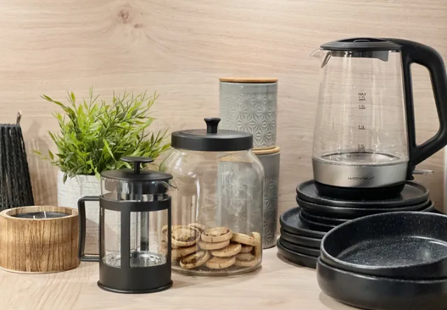 Een gezellig keukentafereel met een glazen waterkoker, een cafetière, een koekjestrommel, plantendecoratie en divers serviesgoed op een houten achtergrond.