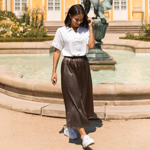 Een vrouw in een wit shirt en een bruine rok loopt langs een fontein omringd door groen, met gele gebouwen op de achtergrond.