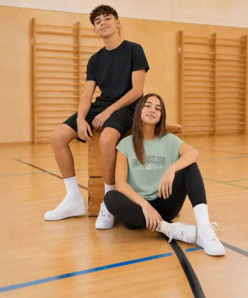 Twee tieners dragen comfortabele sportkleding en zitten in een gymzaal op en naast een springkast. De outfits zijn ideaal voor de gymles en vrije tijd.