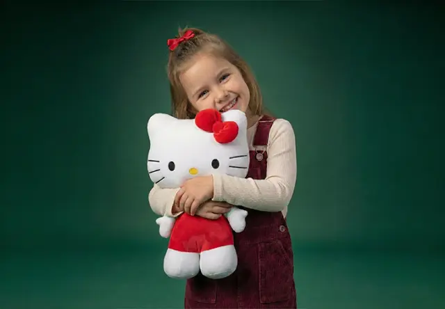 Een kind in een bordeauxkleurige outfit omhelst een grote Hello Kitty-knuffel tegen een groene achtergrond.