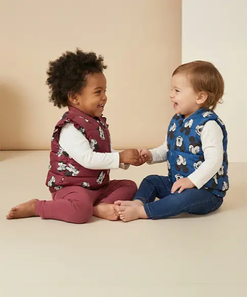 Twee peuters zitten voor een lichte achtergrond, dragen kleurrijke vesten, houden elkaars hand vast en lachen elkaar toe.