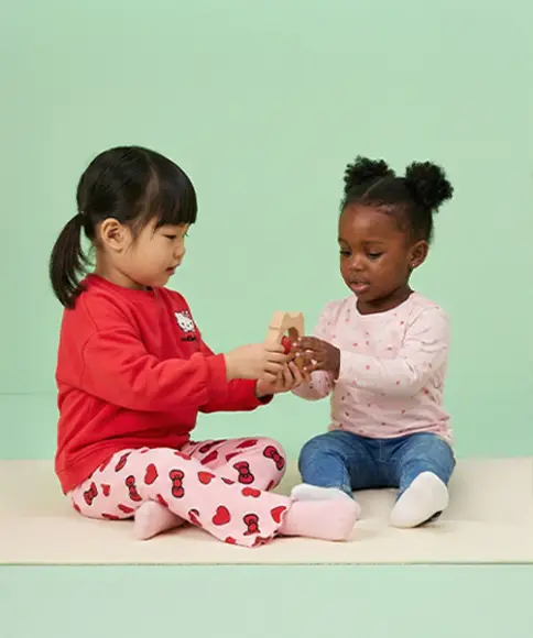 Twee kleine kinderen zitten op een zachte ondergrond en spelen vrolijk samen, terwijl ze kleurrijk speelgoed vasthouden tegen een pastelgroene achtergrond.