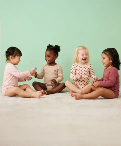 Een groep van vier peuters zit op een lichte vloer en speelt samen met speelgoed voor een effen pastelgroene achtergrond.