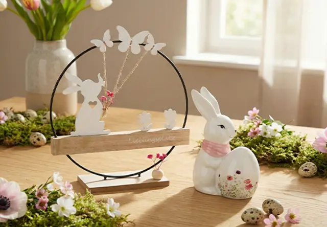 Decoratieve lentescène met een konijnenfiguur met een ei, omringd door vlinders, bloemen en groen op een houten tafel.
