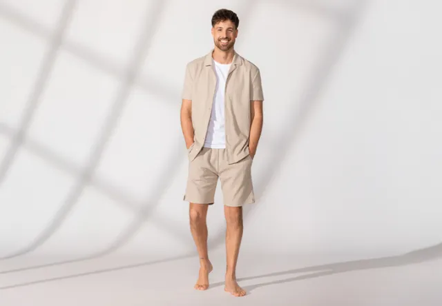Een man staat blootsvoets, draagt een beige overhemd met korte mouwen en bijpassende shorts, met zijn handen nonchalant in de zakken – tegen een lichte achtergrond.