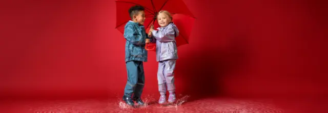 Twee kinderen in felgekleurde regenjassen spelen vrolijk in plassen terwijl ze een felrode paraplu delen.
