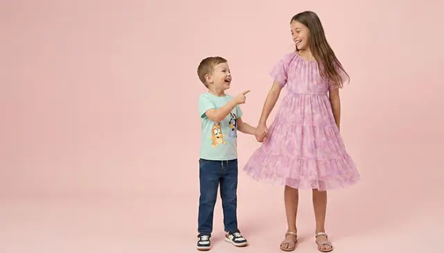 Een jongetje in een lichtblauw shirt en spijkerbroek wijst naar een meisje in een roze jurk met ruches. Ze houden elkaar bij de hand tegen een zachtroze achtergrond.