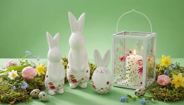 Drie keramische konijntjes, een ei met patroon en een lantaarn met een kaars, omringd door kleurrijke bloemen en paaseieren op een groene achtergrond.