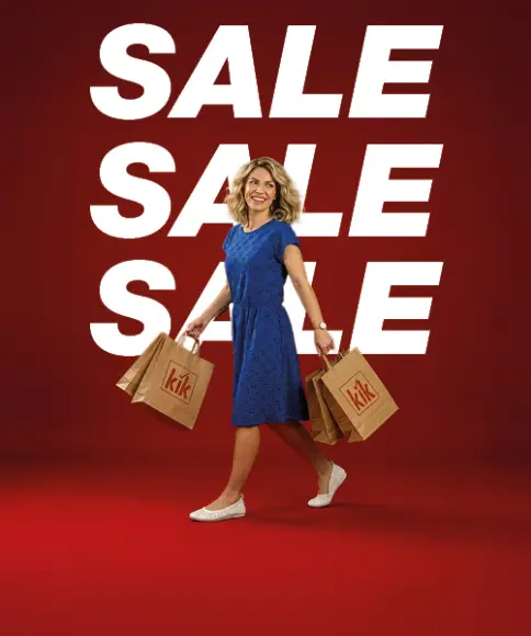 Een vrouw in een blauwe jurk draagt meerdere boodschappentassen met KiK‑logo en loopt voor een grote witte „SALE”-tekst op een rode achtergrond.