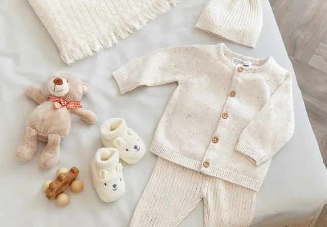 Gezellige babyuitzet met een gebreid vest, bijpassende broek, een muts, slofjes, een teddybeer en houten speelgoed op een zachte deken.