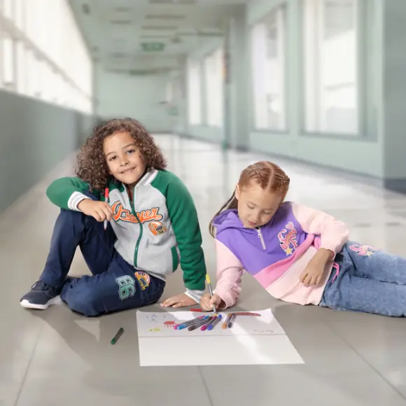 Twee kinderen zitten op een tegelvloer en tekenen met gekleurde stiften op een groot vel papier.