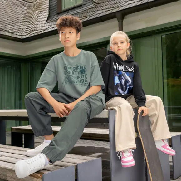 Een jongen in een oversized groen T-shirt en groene broek zit op een houten bank naast een meisje in een zwarte hoodie en beige broek, met een skateboard in haar hand.