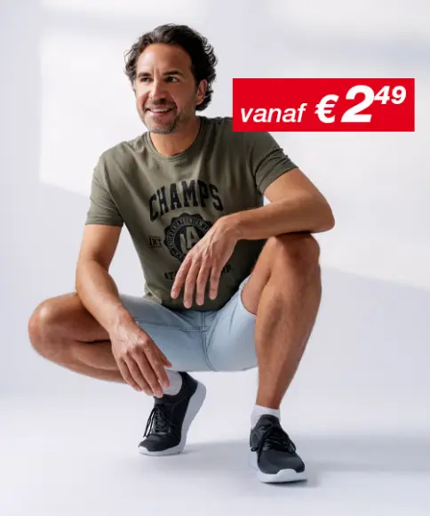 Een man in een nonchalante houding, gekleed in een groen T-shirt en lichtblauwe korte broek, met zwarte sportschoenen, voor een zachte achtergrond.