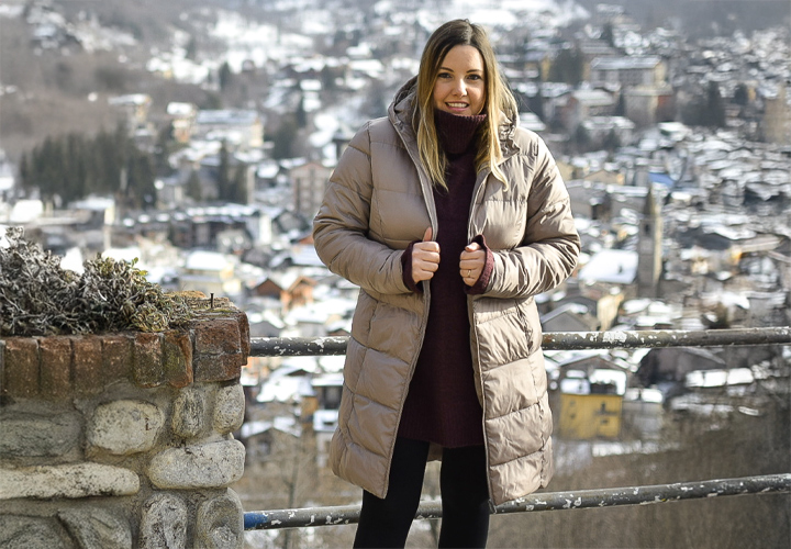 Een vrouw in een stijlvolle beige gewatteerde jas staat bij een stenen reling en kijkt uit over een besneeuwd, pittoresk bergdorp.