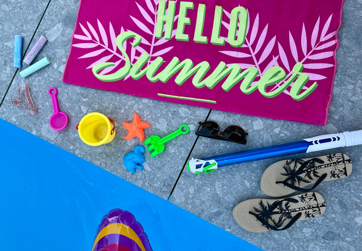 Een levendig zomers tafereel met een roze handdoek met de tekst ‘Hello Summer’, strandspeelgoed, een zonnebril en teenslippers naast een zwembad.