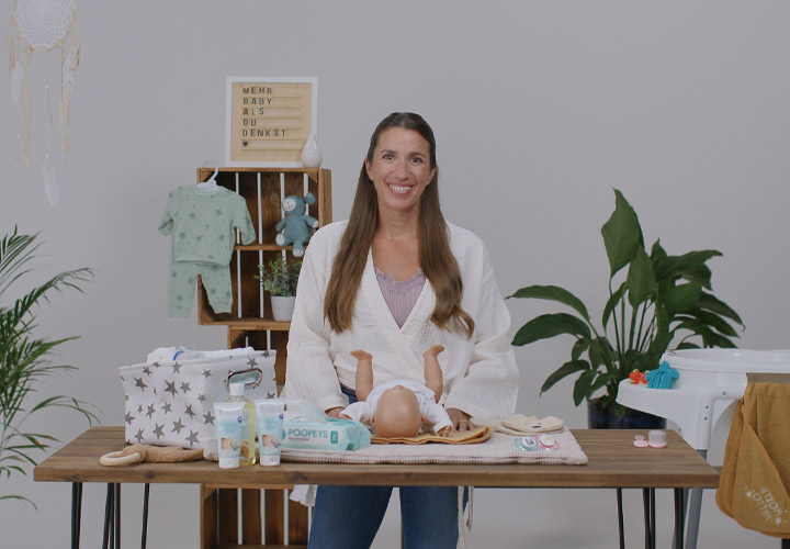 Een vrouw staat aan een houten tafel met babyverzorgingsproducten, kleding en speelgoed, omringd door groen in een lichte omgeving. Een vrouw staat aan een houten tafel met babyverzorgingsproducten, kleding en speelgoed, omringd door groen in een lichte omgeving.