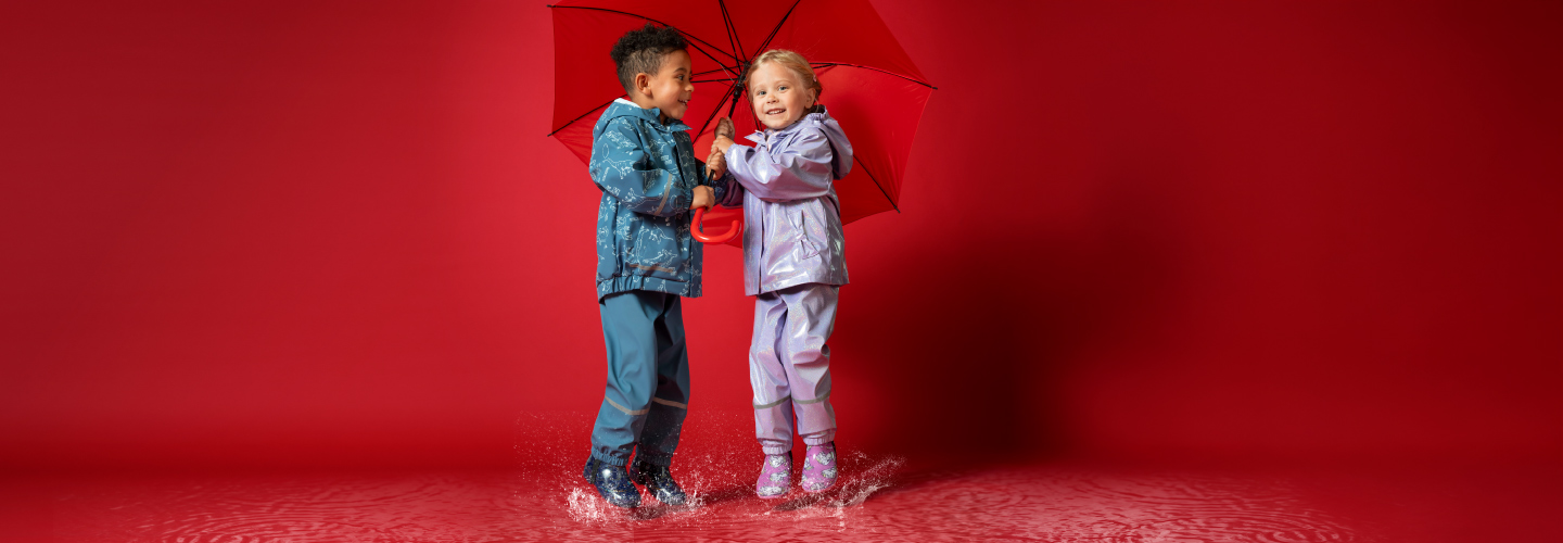Een kind speelt in de regenachtige natuur en draagt een beige regenjas met kattenmotieven en de bijpassende bruine broek.