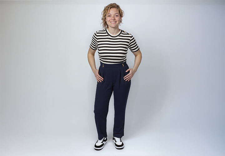 Een persoon staat zelfverzekerd in een gestreept shirt en een marineblauwe high-waisted broek, gecombineerd met zwart-witte schoenen, tegen een egale achtergrond.