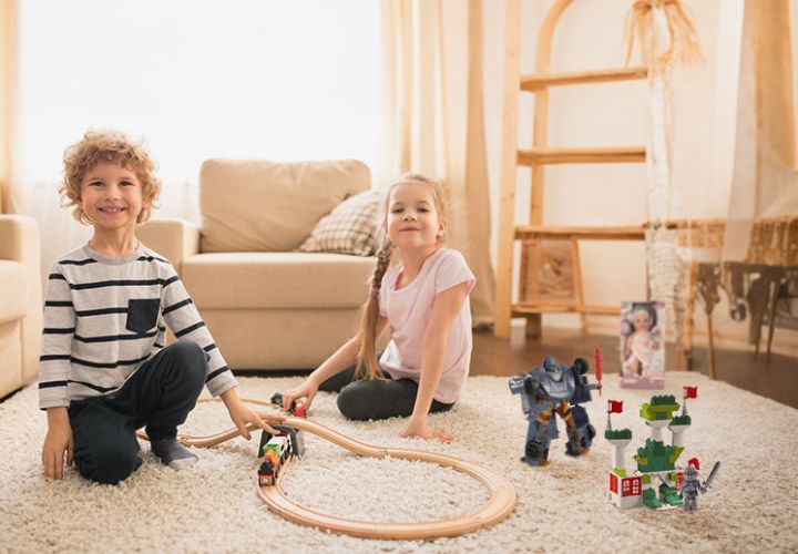 Twee kinderen spelen op een tapijtvloer met een houten trein, een speelgoedrobot en een bouwset, omringd door een gezellig woonkamer.