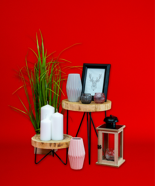 Decoratieve opstelling met houten plantenstandaards, kaarsen, vazen, een fotolijst en een lantaarn tegen een felrode achtergrond.