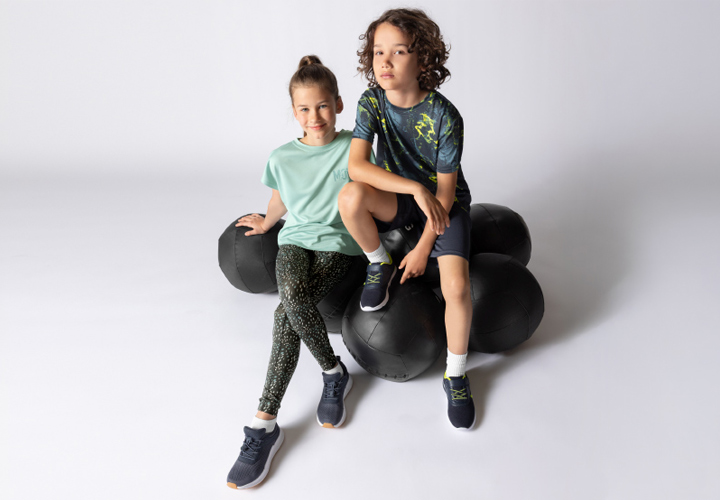 Twee kinderen zitten op een groep zwarte fitnessballen, gekleed in kleurrijke sportkleding en sportschoenen, tegen een neutrale achtergrond.