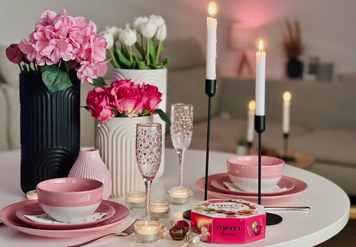 Een prachtig gedekte dinertafel met roze borden, kaarsen, elegant glaswerk en een doos Merci-chocolade, omringd door verse bloemen.