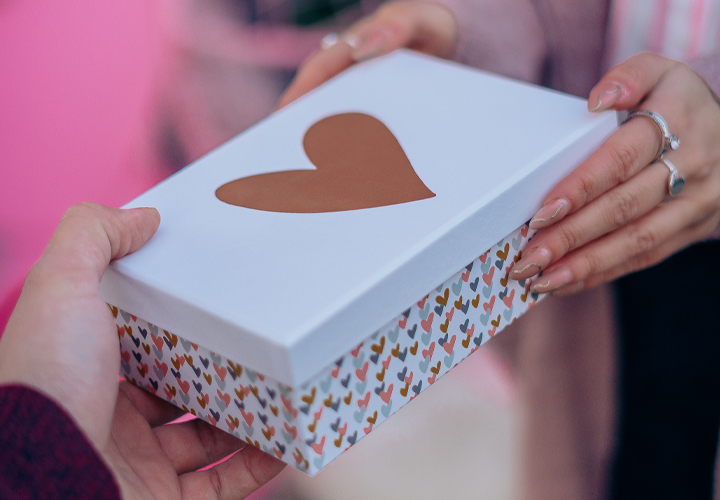 Een persoon overhandigt een decoratieve geschenkdoos, versierd met een hart en kleurrijke patronen.